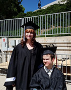 young woman stands next to a young man in a wheelchair both wearing graduation cap and gown